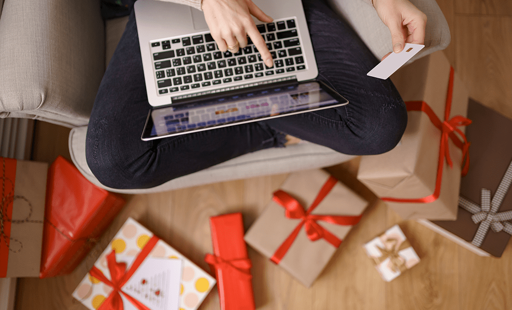 Black Friday. Pessoa sentada numa poltrona com as pernas cruzadas e um portátil no colo. Está a realizar uma compra online, segurando um cartão de crédito na mão direita. Ao redor da poltrona, no chão, há vários presentes embrulhados com cores diferentes. O ambiente sugere uma atmosfera de compras de fim de ano, possivelmente associada à Black Friday e ao Natal.