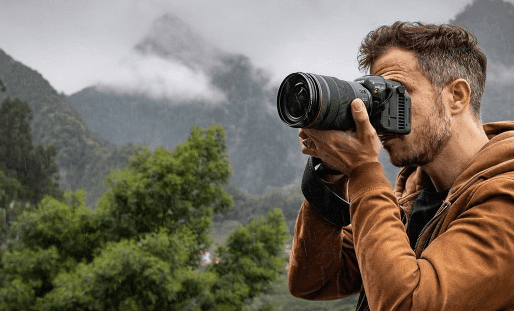 Descrição. Fotógrafo a utilizar a câmara digital Canon EOS R5, de cor preta, para capturar imagens e vídeos de uma bela paisagem em 8K.