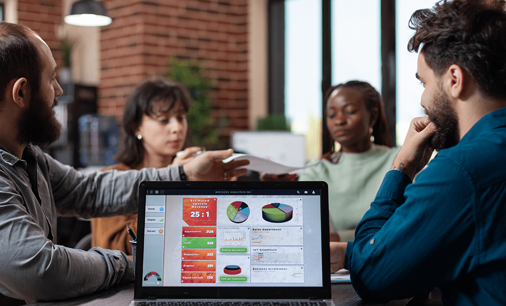 Descrições. Quatro pessoas de diferentes etnias numa reunião de negócios, a discutir gráficos exibidos no ecrã de um computador portátil.
