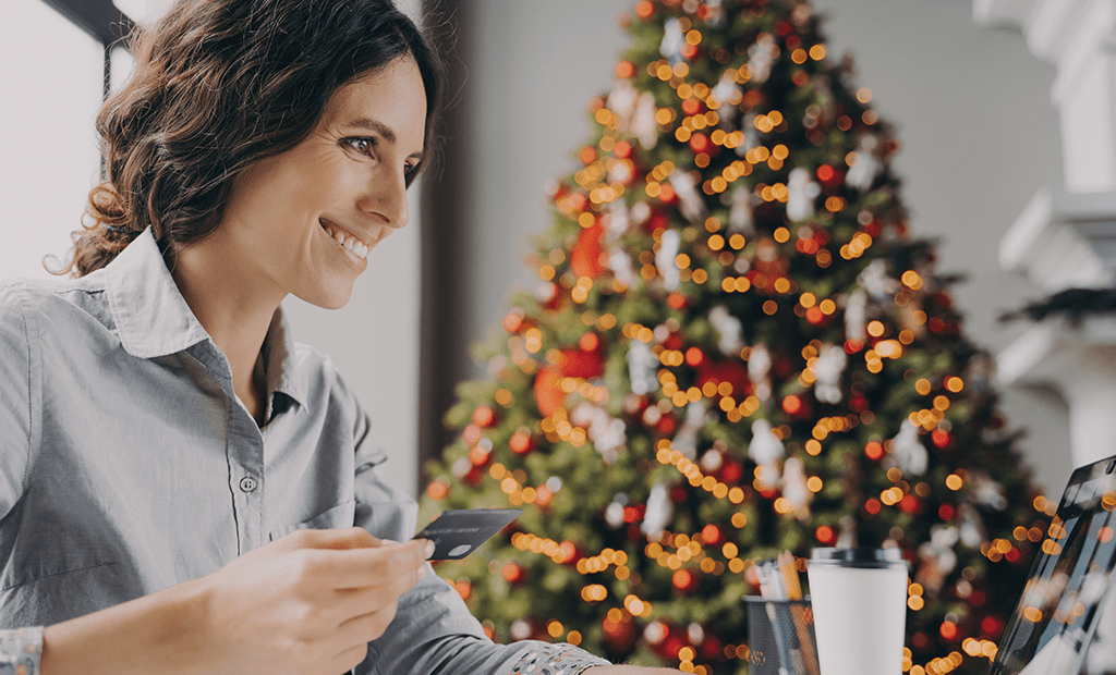 Mulher sorridente sentada em frente a um computador, a segurar um cartão de crédito enquanto realiza compras online. Ao fundo, uma árvore de Natal decorada com luzes e enfeites, criando um ambiente festivo e acolhedor.