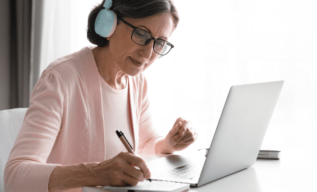Mulher madura com auscultadores, a utilizar um computador portátil e a tomar notas, simbolizando o acesso inclusivo à educação online para todas pessoas.