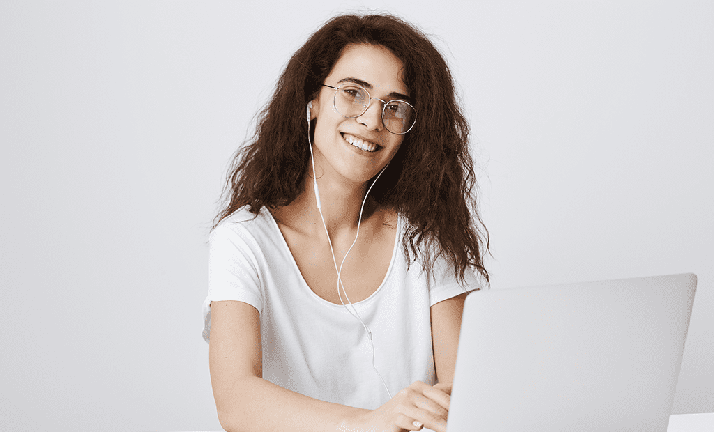 Mulher jovem com cabelo castanho ondulado, usando óculos e auscultadores brancos, a sorrir enquanto trabalha num computador portátil. O fundo é branco e minimalista.