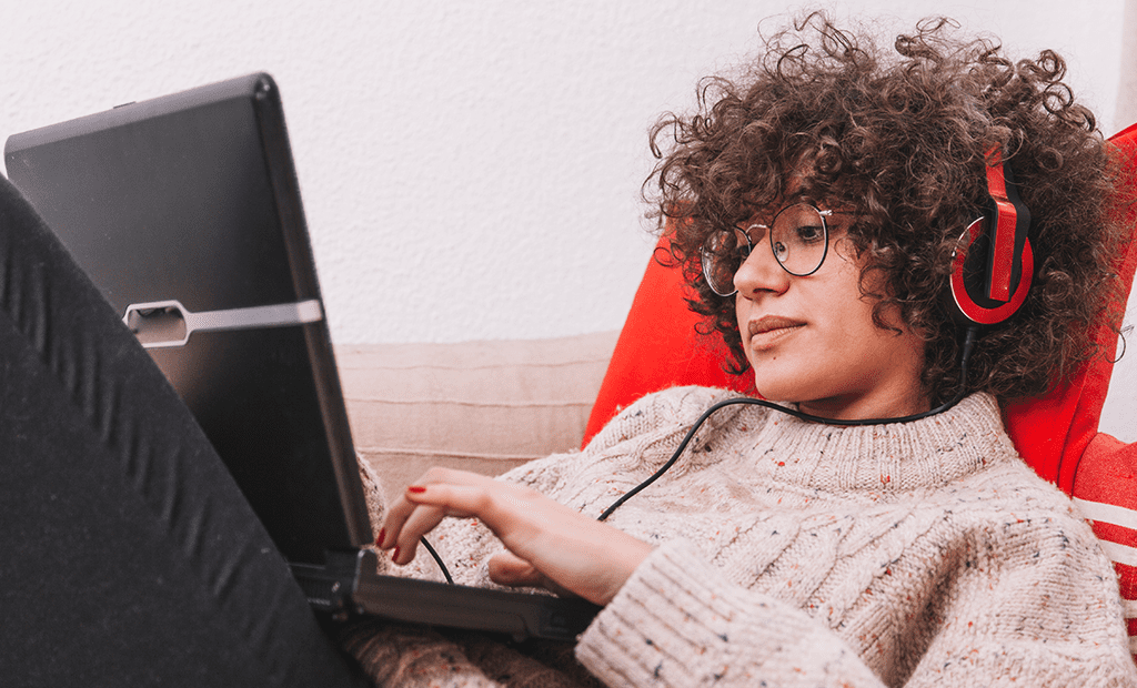 Leitor de ecrã. Mulher com cabelo encaracolado e óculos, sentada num sofá, utiliza um computador portátil com auscultadores vermelhos, simbolizando o uso de tecnologias assistivas auditivas como leitores de ecrã.