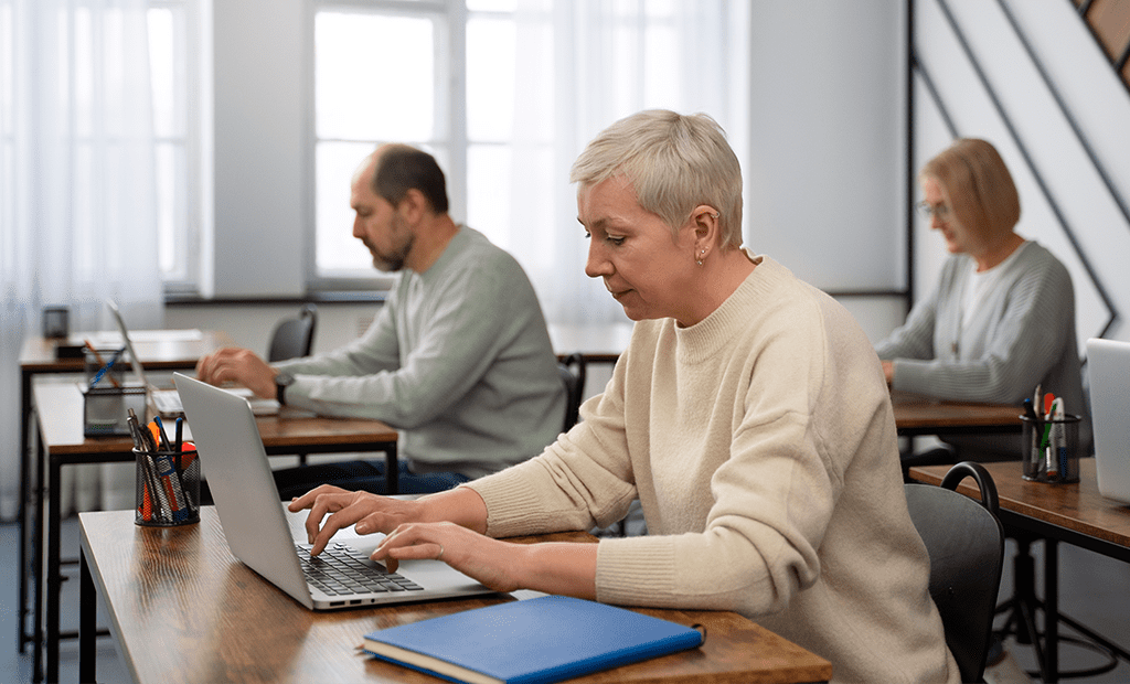 Leitor de ecrã. Três adultos sentados em sala de formação, cada um a utilizar um computador portátil. Em primeiro plano, uma mulher de cabelo curto e grisalho digita concentrada, representando aprendizagem digital acessível.