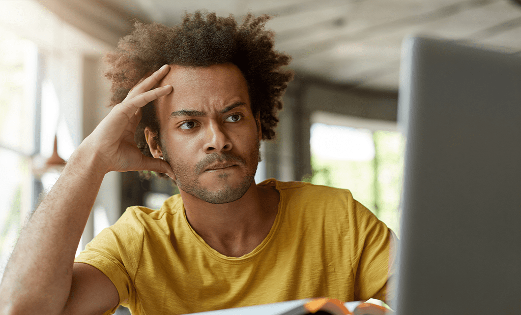 Leitor de ecrã. Homem com expressão concentrada e ligeiramente frustrada, apoiando a cabeça na mão enquanto olha para um ecrã de computador, sugerindo dificuldades de navegação ou de acessibilidade digital.