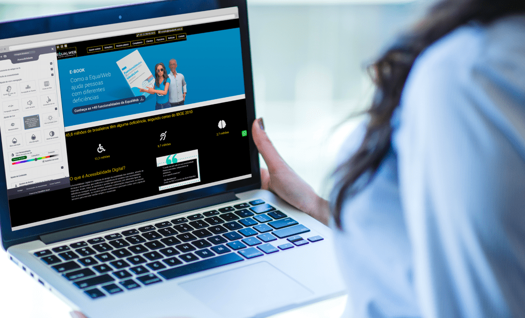 Leitor de ecrã. Mulher segura um portátil com o site da EqualWeb aberto em modo de alto contraste e o menu de acessibilidade visível, ilustrando a personalização da experiência digital para utilizadores com deficiência visual.